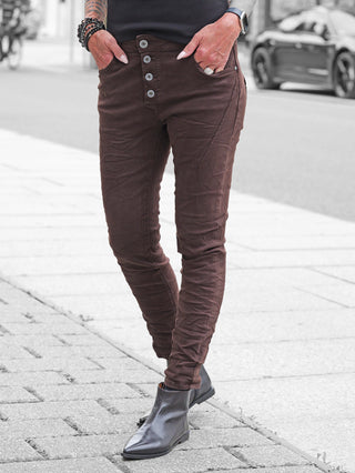 Person trägt OFF#DLY Color Jeans Imelda schoko, schlanke Passform, Knopfleiste sichtbar. Hose in Stiefel gesteckt, Hand in Tasche, Schmuck erkennbar, ideal für stilvolle Alltags- oder Casual-Looks.