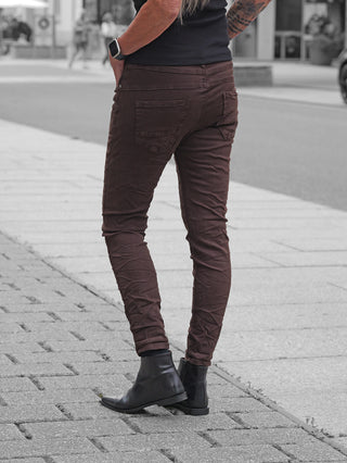 Person trägt OFF#DLY Color Jeans Imelda schoko, schmal geschnittene Hose aus leichtem Stretch-Material, kombiniert mit schwarzen Lederstiefeletten. Hände in Taschen, eine Smartwatch am Handgelenk.