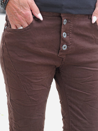 Nahaufnahme der unteren Körperhälfte, die OFF#DLY Color Jeans Imelda schoko zeigt: dunkle braune Hose mit Drei-Knopf-Verschluss, Hände in den vorderen Taschen, ergänzt durch Ring und Armbänder.