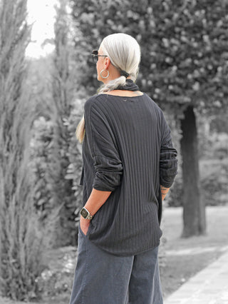 Eine Person trägt das OFF#DLY Longsleeve Sigrid black, ein geripptes, lockeres schwarzes Oberteil mit hochgeschobenen Ärmeln, kombiniert mit weiten grauen Hosen, und steht auf einem Gartenweg.