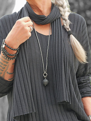 Person in einem charcoal-grauen OFF#DLY Longsleeve Sigrid mit integriertem Schal, silbernem Kettenanhänger, schwarzen Armbändern, silbernem Ring, seitlichem Zopf und tätowiertem Unterarm sichtbar.