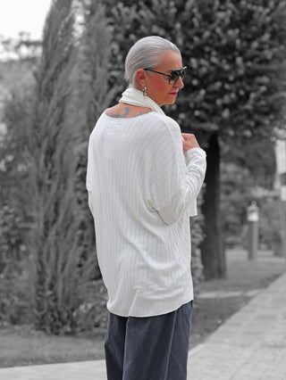 Ältere Frau trägt ein OFF#DLY Longsleeve Sigrid offwhite mit lockerem Schnitt und zugehörigem Schal, aus Rippenstrick, während sie auf einem Gehweg mit gepflegten Hecken spaziert.
