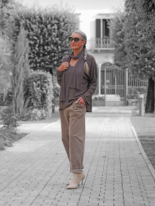 Reife Frau in OFF#DLY Longsleeve Sigrid schoko, elegant gerippter Stoff mit passendem Schal, trägt große Sonnenbrille, lange graue Haarzopf, steht vor Haus auf gepflastertem Gartenweg.
