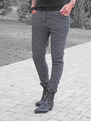 OFF#DLY Muster-Pant Saga grey/black: Person trägt eng geschnittene Hose mit geometrischem Muster, kombiniert mit schwarzen bestickten Lederstiefeletten. Hände in den Taschen, zeigen Ringe, Armbänder und tätowierten Unterarm.