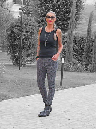 Eine Frau trägt die OFF#DLY Muster-Pant Saga grey/black: gemusterte, schmal geschnittene Hose, kombiniert mit einem schwarzen Tanktop und Stiefeletten.