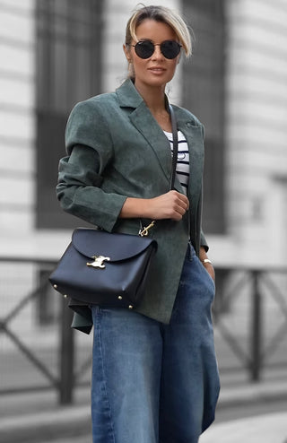 Frau in urbanem Setting trägt Gitta Banko Blazer Amy khaki, kombiniert mit gestreiftem Shirt und blauer Jeans, abgerundet durch schwarzen Ledersatchel.