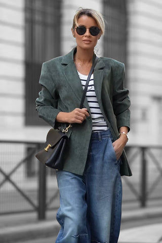 Frau trägt den Gitta Banko Blazer Amy khaki, locker geschnitten, über gestreiftem Shirt und blauen Jeans, während sie eine schwarze Tasche hält, im urbanen Umfeld spazierend.