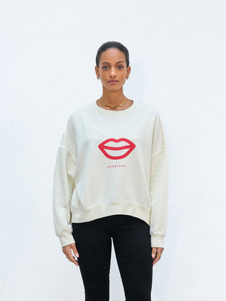 Eine Frau trägt das Outryght Bobbi L white dove Sweatshirt mit einem auffälligen roten Lippen-Print, kombiniert mit schmalen schwarzen Hosen. Sie steht vor einem neutralen Hintergrund, minimalistisch geschmückt.