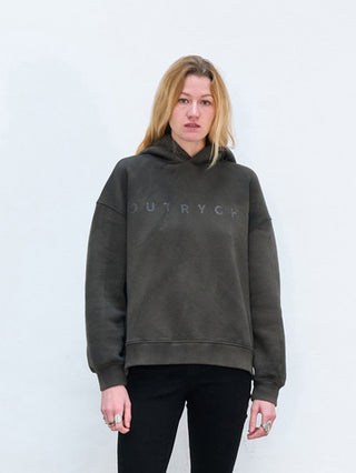 Eine junge Person trägt den Outryght Sweater military, ein dunkel oversized Hoodie mit einem dezenten Logo auf der Brust, kombiniert mit dunklen Hosen und mehreren Ringen an den Fingern.