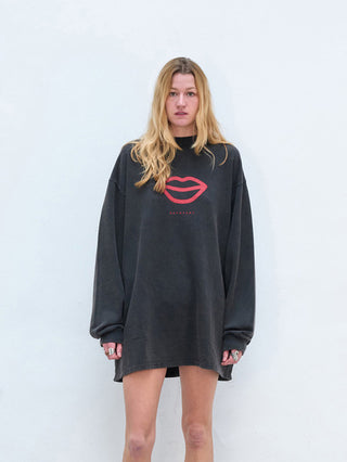 Person trägt das Outryght Tee Dress L in verwaschenem Schwarz, ein übergroßes Sweatshirt mit rotem Lippenmotiv, das bis zu den Oberschenkeln reicht. Lange blonde Haare, neutraler Gesichtsausdruck.