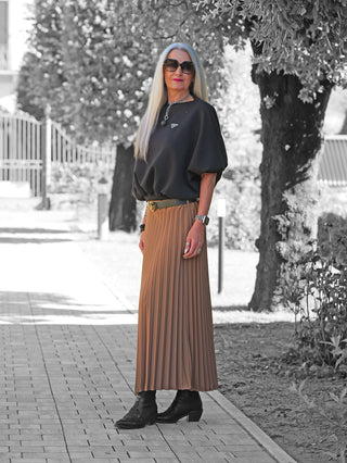 Frau trägt OFF#DLY Plissee Rock camel, einen eleganten, hoch taillierten Faltenrock mit Gürtel, kombiniert mit schwarzen Stiefeln und Accessoires, auf einem baumgesäumten Weg.