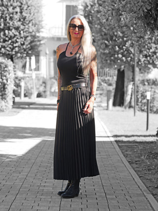 Frau im Garten trägt den OFF#DLY Plissee Rock black: eleganter, knitterfreier Plissee-Rock mit Doppel-G Gürtel, perfekt für vielseitige Stylings. Kombiniert mit schwarzem Top, Stiefeletten und Accessoires.