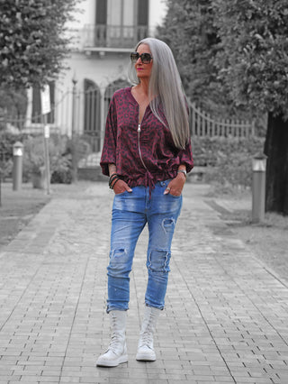 Frau trägt die OFF#DLY Leo Jacke Hedda bordeaux, ein modisches Statement-Piece mit Leo-Muster, kombiniert mit zerrissenen Jeans und weißen Stiefeln, in urbanem Parksetting.
