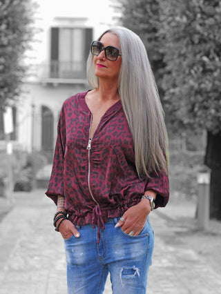 Person mit grauem Haar trägt die OFF#DLY Leo Jacke Hedda bordeaux mit Leopardenmuster, kombiniert mit hellblauen Jeans, Schmuckaccessoires, und blickt in die Ferne.