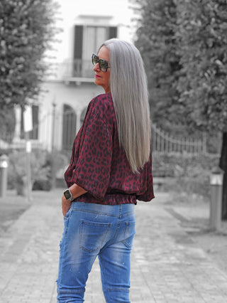 Person mit langen, silber-grauen Haaren trägt die OFF#DLY Leo Jacke Hedda bordeaux, erkennbar am lockeren Schnitt und animalischen Muster, perfekt kombiniert mit Jeans auf einem gepflasterten Weg.