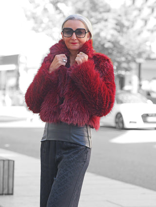 Eine reife Frau trägt die OFF#DLY Felljacke Luisa bordeaux, mit hochgezogenem Kragen. Die Jacke aus kuscheligem Kunstpelz ergänzt ihr stilvolles Outfit mit dunkler Hose und Leder-Top.
