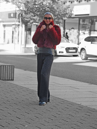 Eine Frau trägt die OFF#DLY Felljacke Luisa bordeaux, ein kurz geschnittener, burgunderfarbener Kunstpelzmantel, kombiniert mit schwarzen Hosen und Sneakers, im urbanen Straßenstil auf einem Gehweg.