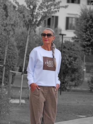 Ältere Frau in OFF#DLY Sweat Latte Choco white, lässigem weißen Sweatshirt mit braunem Late chocolate-Print, kombiniert mit brauner Cordhose, stehend auf gepflastertem Platz im Freien.