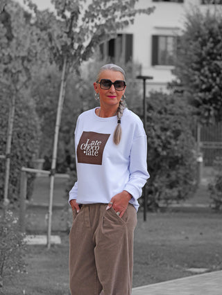 OFF#DLY Sweat Latte Choco white: Frau trägt weißes Sweatshirt mit Late choco·late-Print, kombiniert mit braunen Cordhosen, in einem parkähnlichen Umfeld. Stilvoll, lässig und vielseitig kombinierbar.