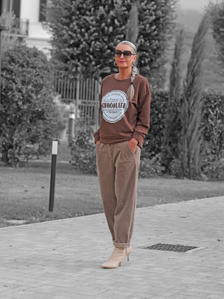 Frau trägt OFF#DLY Sweat Chocolate braun mit rundem Logo-Aufdruck, kombiniert mit lässiger Cordhose und Stiefeletten. Perfekt für einen entspannten, stylischen Look im Garten oder Hof.