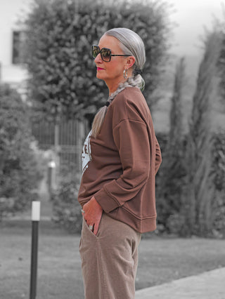 Ältere Frau im Garten trägt das OFF#DLY Sweat Chocolate braun, erkennbar am weißen Frontprint, kombiniert mit hellbrauner Cordhose und Sonnenbrille, Hand lässig in der Tasche.