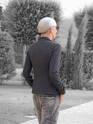 Person von hinten in einem OFF#DLY Blazer Tiziana black mit schmalem Schnitt und Ein-Knopf-Design, kombiniert mit lässiger Cargo-Hose, steht auf einem gepflasterten Weg zwischen gepflegten Hecken.