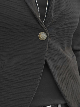 Nahaufnahme des OFF#DLY Blazer Tiziana schwarz mit Waffelstruktur, verziert mit einem goldenen Knopf. Darunter ist ein schwarzes Oberteil mit sichtbarem silbernem Reißverschluss zu erkennen.