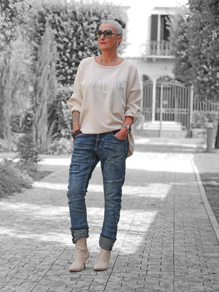 Eine stilvolle Frau trägt die OFF#DLY Denim Ilaria darkblue, eine lässig-edgy Jeans mit Destroy-Details und sportivem Schnitt, kombiniert mit einem cremefarbenen VOGUE-Sweatshirt und beigen Stiefeletten.