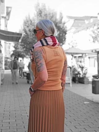 Frau in schwarz-weißem Hintergrund trägt das OFF#DLY Top Neve camel mit Smiley-Patch, kombiniert mit einem langen Rock und einem gestreiften Schal, betont durch Tattoos und eine moderne Smartwatch.