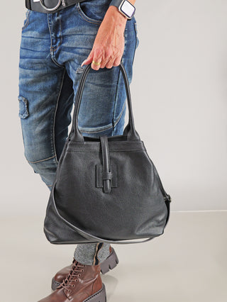Person hält die OFF#DLY Leder Tasche black, eine schwarze Ledertasche mit Henkeln, kombiniert mit blauen Jeans und braunen Stiefeln. Sichtbar sind ein Karabiner-Verschluss und ein langer Trageriemen.