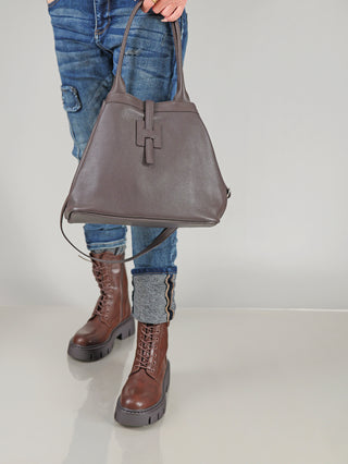 Eine Person trägt eine OFF#DLY Leder Tasche schoko aus grauem, genarbtem Leder mit zwei Henkeln und einem tab-and-loop Verschluss, passend zu braunen Lederstiefeln und blauen Jeans.