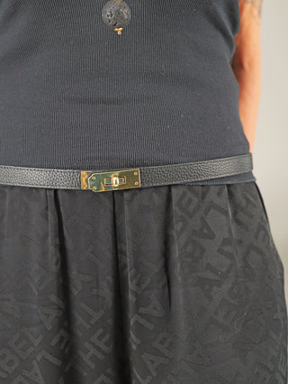 Nahaufnahme eines Outfits mit dem OFF#DLY Gürtel Leder black, einem schlanken Ledergürtel mit goldfarbener Drehverschluss-Schnalle, kombiniert mit einem schwarzen, gerippten Tanktop und einem schwarzen Rock mit erhabenen Buchstaben.
