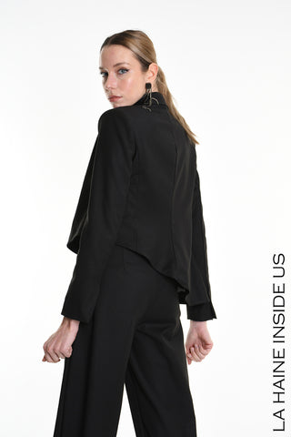 Modell trägt La Haine Jacket black, eine asymmetrische, fließende, gefütterte Jacke mit Knopfverschluss und Innenschultern, kombiniert mit passenden weiten Hosen. Statement-Ohrring ergänzt den Look.