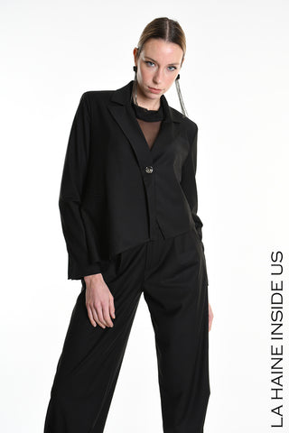 Model trägt La Haine Jacket black, eine asymmetrische, leicht übergroße schwarze Jacke mit Knopfverschluss. Kombiniert mit schwarzer Hose und langem Ohrring, vor weißem Hintergrund stehend.