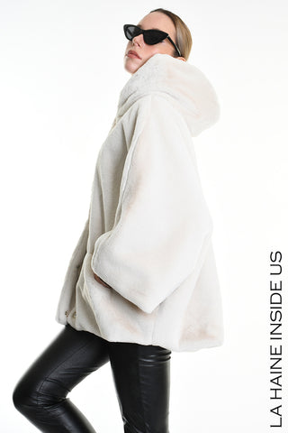Model trägt die La Haine Jacket white, eine voluminöse, off-white Kunstfell-Sportjacke mit Kapuze und Knopfverschluss, kombiniert mit schwarzen Lederleggings und cat-eye Sonnenbrille, vor weißem Hintergrund.