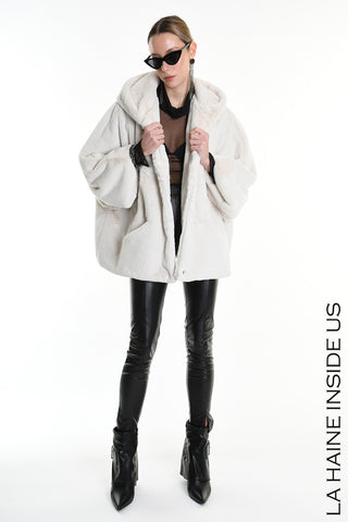 Model trägt den La Haine Jacket white, eine oversized Kunstfelljacke mit Kapuze und Knopfverschluss, über einem transparenten schwarzen Top, kombiniert mit schwarzen Lederleggings und spitzen Stiefeletten.