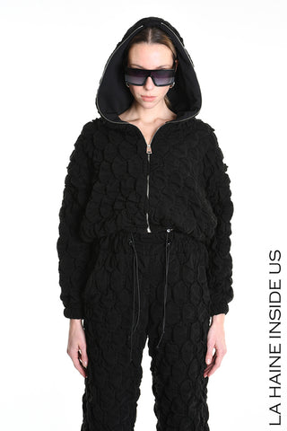 Person trägt die La Haine Bomber Jacket black, eine schwarze, geprägte Kapuzenjacke mit Reißverschluss und passenden Jogginghosen. Oversize-Sonnenbrille vervollständigt den Look. Markentext LA HAINE INSIDE US sichtbar.