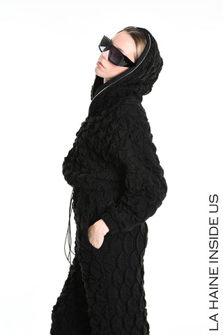 Model in Profilansicht trägt La Haine Bomber Jacket black, eine texturierte, gesteppte Kapuzenjacke mit Reißverschluss und elastischen Bündchen, kombiniert mit passenden Hosen und übergroßen schwarzen Sonnenbrillen.