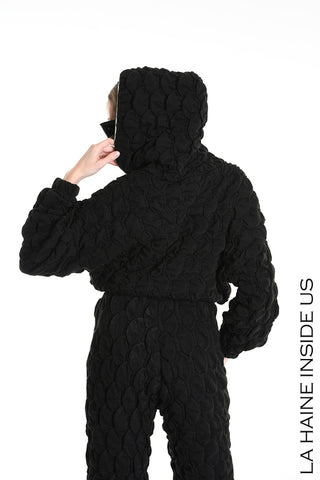 Person von hinten in einem schwarzen, gesteppten Einteiler mit Kapuze, trägt die La Haine Bomber Jacket black. Strukturierte, gepolsterte Oberfläche, linke Hand zum Gesicht erhoben, Text rechts: “LA HAINE INSIDE US.”