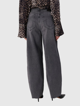 Model von hinten in True Religion Ruby Volume Jeans, hochgeschnitten, weit, mit offenem Saum, Patch-Taschen. Kombiniert mit eingesteckter Leopardenbluse und schwarzen Stilettos, vor grauem Studiohintergrund.