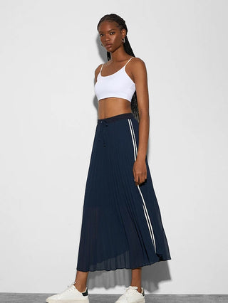 Junge Frau im Profil trägt den BSB Skirt French Navy, einen navyblauen, plissierten Midi-Rock mit seitlichen weißen Streifen, kombiniert mit einem weißen Crop-Top und weißen Sneakers.