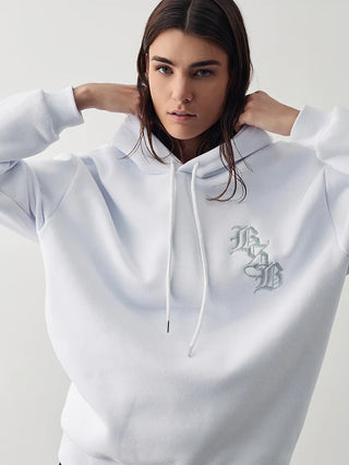 Eine junge Frau trägt die BSB Bluse 210084 white, ein weißes Kapuzen-Sweatshirt mit einem dezentem, gotischen Monogramm auf der Brust, und hebt den Hoodie mit beiden Händen an.
