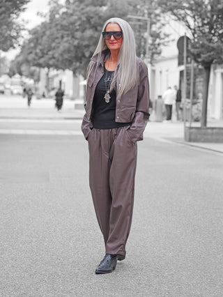 Eine stilvolle Frau trägt die BSB Hose 212003 chocolate, eine locker geschnittene, hochtaillierte Kunstlederhose in Braun mit seitlichen Eingrifftaschen und elastischem Bund, kombiniert mit schwarzem Tanktop und Hamsa-Anhänger.