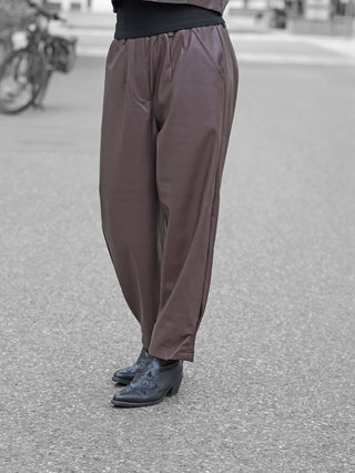 BSB Hose 212003 chocolate: Locker sitzende, schokoladenbraune Kunstlederhose mit leichtem Bundfalten-Design, kombiniert mit schwarzen, dezent bestickten Lederstiefeletten auf gepflasterter Straße.