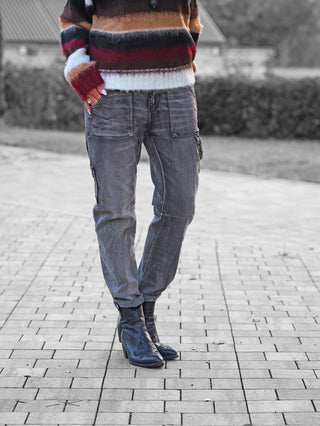 Person trägt OFF#DLY Denim Beatrice black washed: graue, locker sitzende Cargo-Jeans mit markanten, aufgesetzten Taschen und destroyed-Style. Kombiniert mit gestreiftem Strickpullover und schwarzen Lederstiefeln.