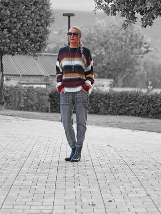 Frau trägt den OFF#DLY Pulli Silvana Stripe black/bordeaux mit auffälligen Multicolor-Streifen und lockerem Schnitt, kombiniert mit Cargo-Jeans und Lederstiefeln, in einem parkähnlichen Umfeld.