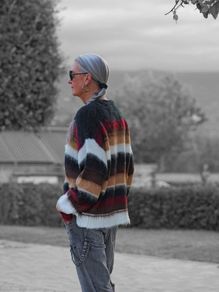 Person im Profil trägt den OFF#DLY Pulli Silvana Stripe black/bordeaux, einen kuscheligen Pullover mit auffälligem Multicolor-Streifenmuster, kombiniert mit grauen Cargo-Jeans und Sonnenbrille.