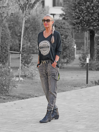Frau mit silbergrauem Zopf trägt OFF#DLY Longsleeve Chocolate black, V-Ausschnitt und Frontprint Chocolate, kombiniert mit grauen Utility-Jeans und schwarzen Lederstiefeletten, in einem Gartenweg stehend.