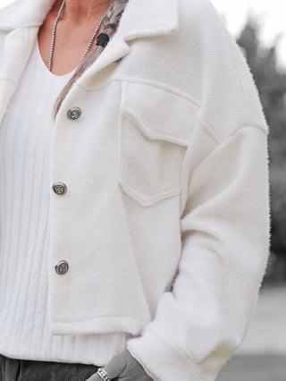 Person trägt die OFF#DLY Jacke Zaira offwhite, leicht verkürzt und strukturiert, mit silbernen Knöpfen und Brusttaschen, über einem weißen Oberteil. Eine Kette und Ringe ergänzen den Look.