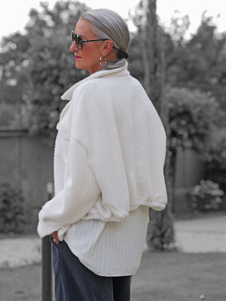Person trägt die OFF#DLY Jacke Zaira offwhite mit kurzem Schnitt, markanten Knöpfen und aufgesetzten Brusttaschen, kombiniert mit dunkler Hose und Sonnenbrille, vor einem Gartenhintergrund.